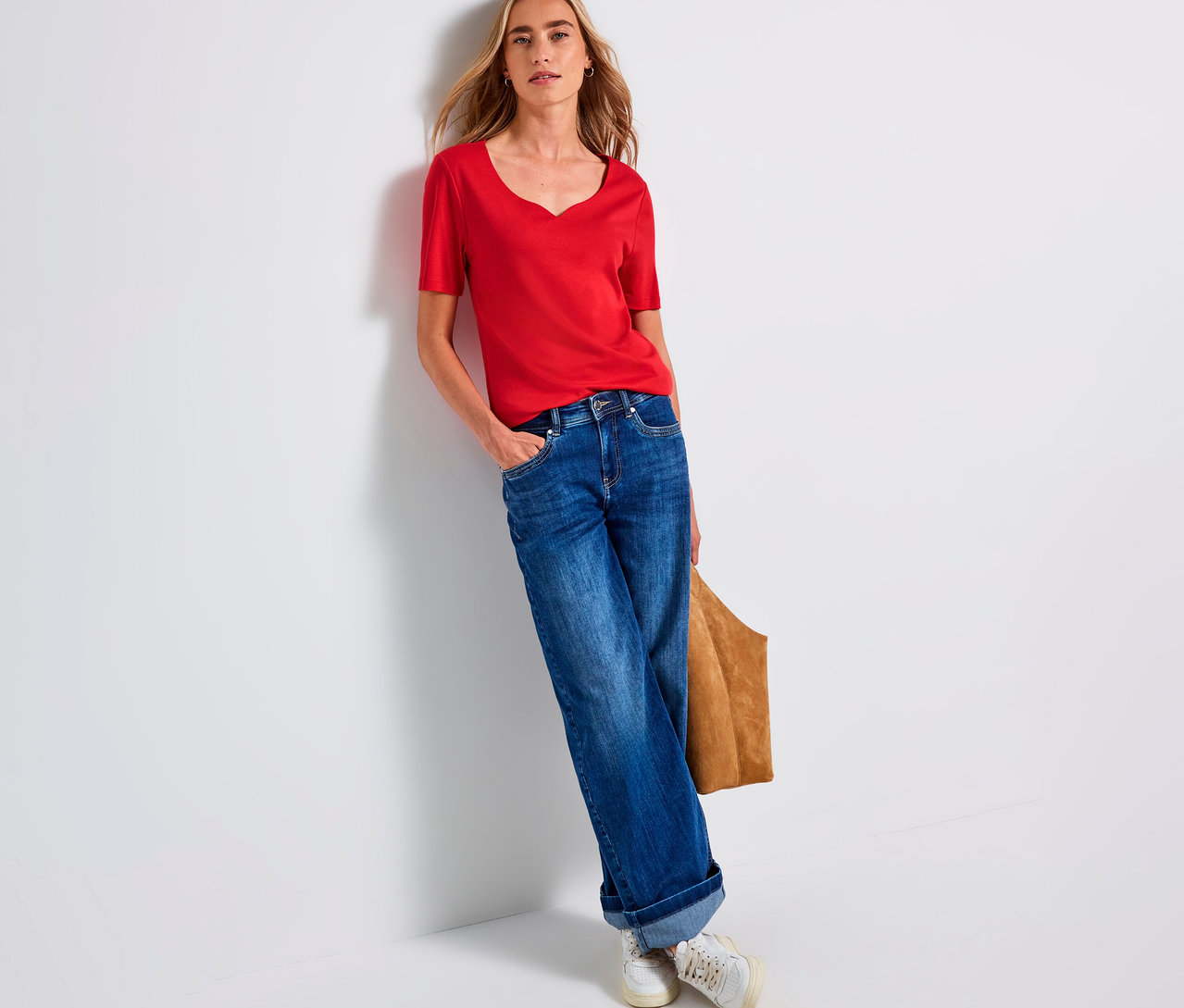 Frau lehnt an einer Wand und trägt ein rotes Street One Shirt mit Herzausschnitt, Jeans und Turnschuhe.