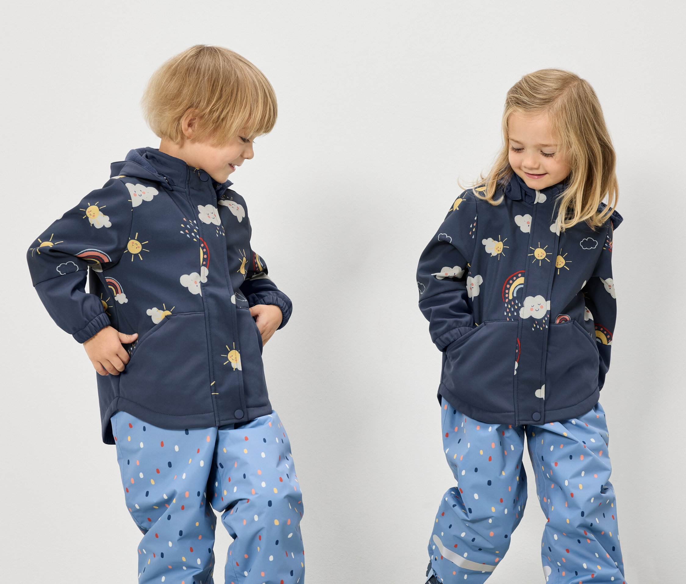 Zwei Kinder mit blauen Jacken mit Regenbogem-, Wolken- und Sonnenmuster und blauen Hosen mit Punktmuster stehen vor einem weißen Hintergrund.