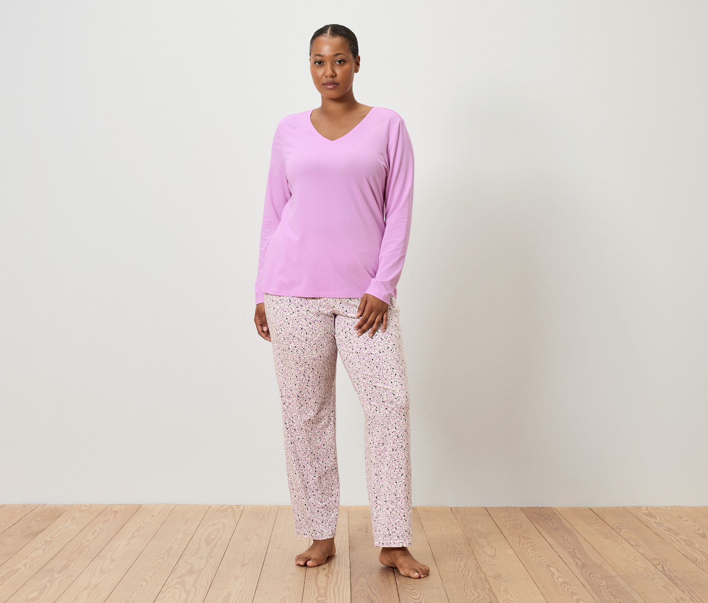 Eine Frau steht barfuß vor einer weißen Wand und trägt ein lila Pyjamaoberteil und eine rosafarbene Pyjamahose mit Blumenmuster.