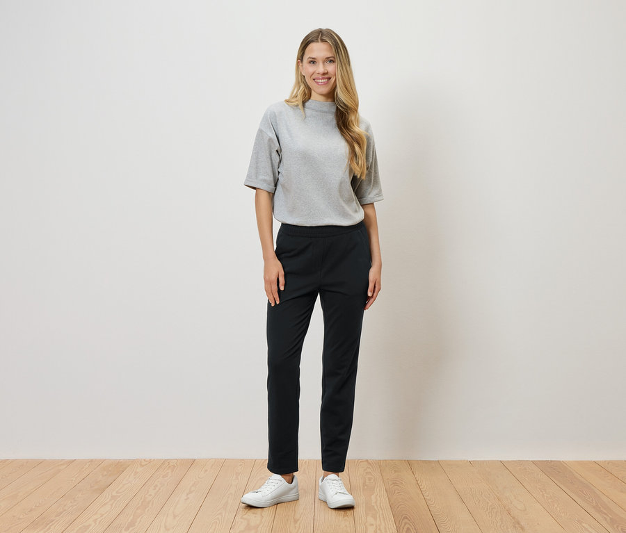 Eine Frau mit blonden Haaren steht vor einer weißen Wand und trägt ein graues T-Shirt, eine schwarze Hose und weiße Turnschuhe.