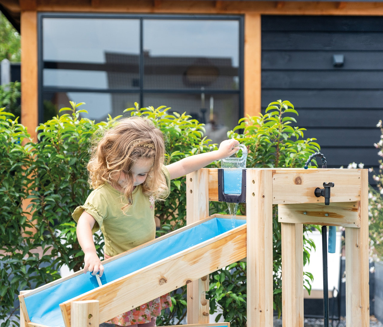 Mädchen gießt Wasser aus einem Krug in eine hölzerne Wasserspielbahn im Garten.
