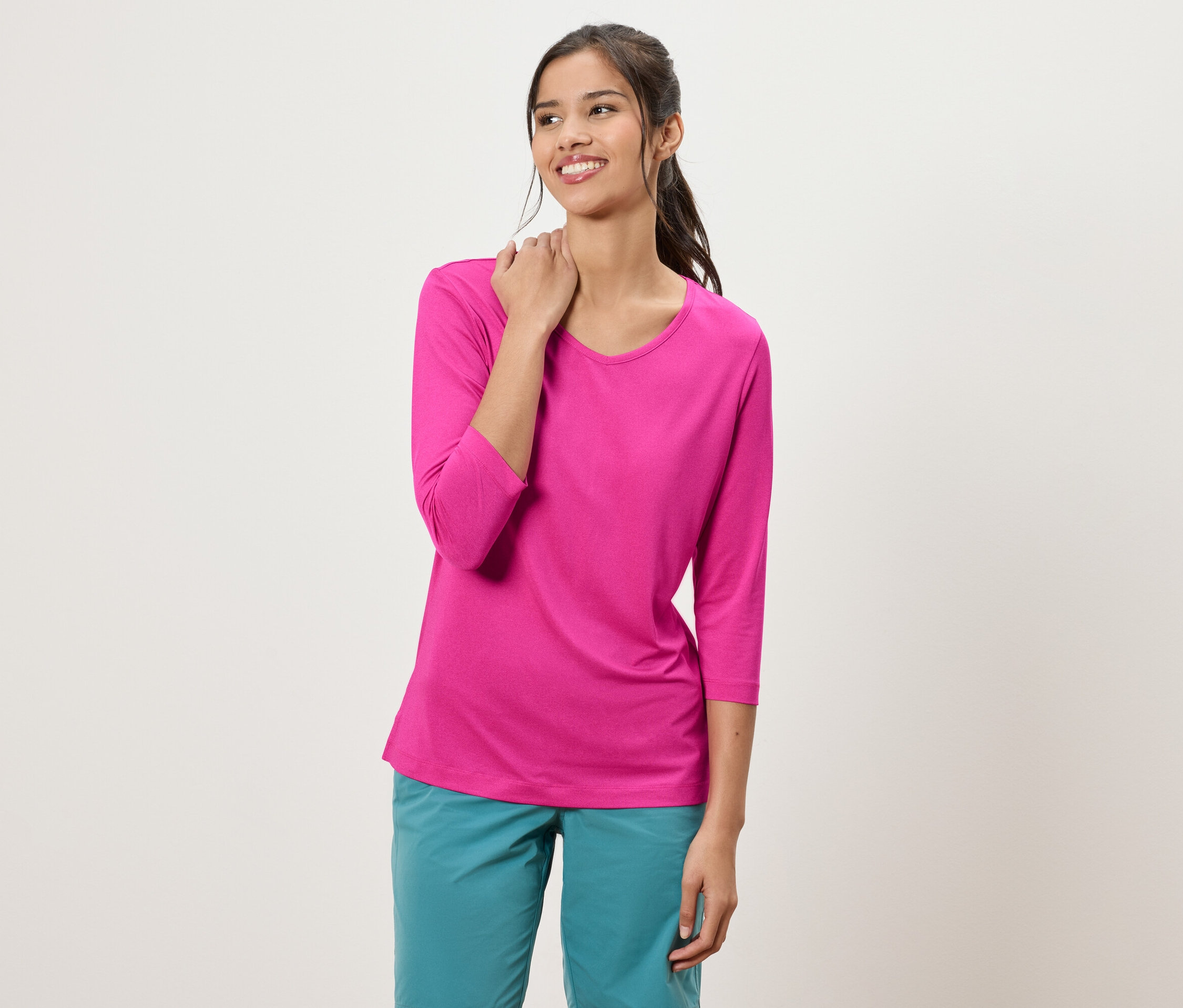 Frau mit Zopf trägt ein pinkes Oberteil mit Dreiviertelärmeln und türkisfarbene Hosen.