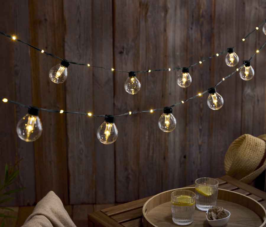 Zwei Reihen Glühbirnen hängen vor einer Holzwand. Auf einem Holztisch stehen zwei Gläser Wasser mit Zitrone und eine Schale mit Nüssen.