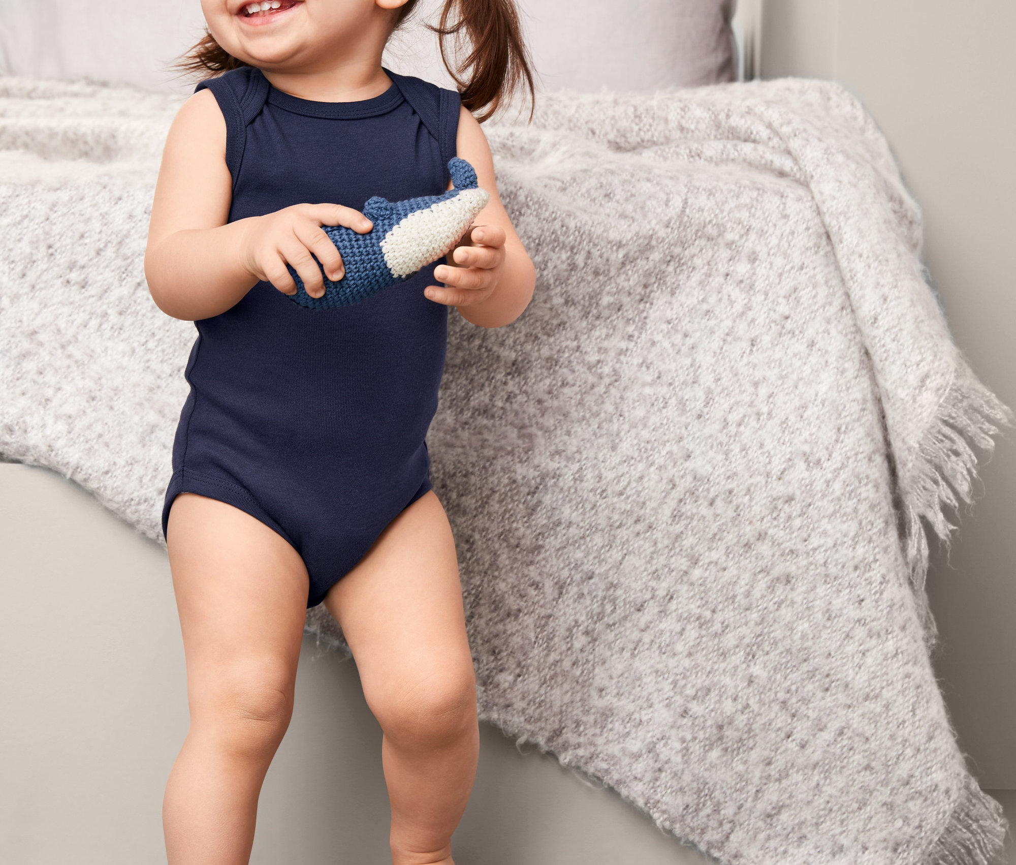 Lächelndes kleines Mädchen in einem marineblauen, ärmellosen Body hält ein blau-weißes Spielzeug.