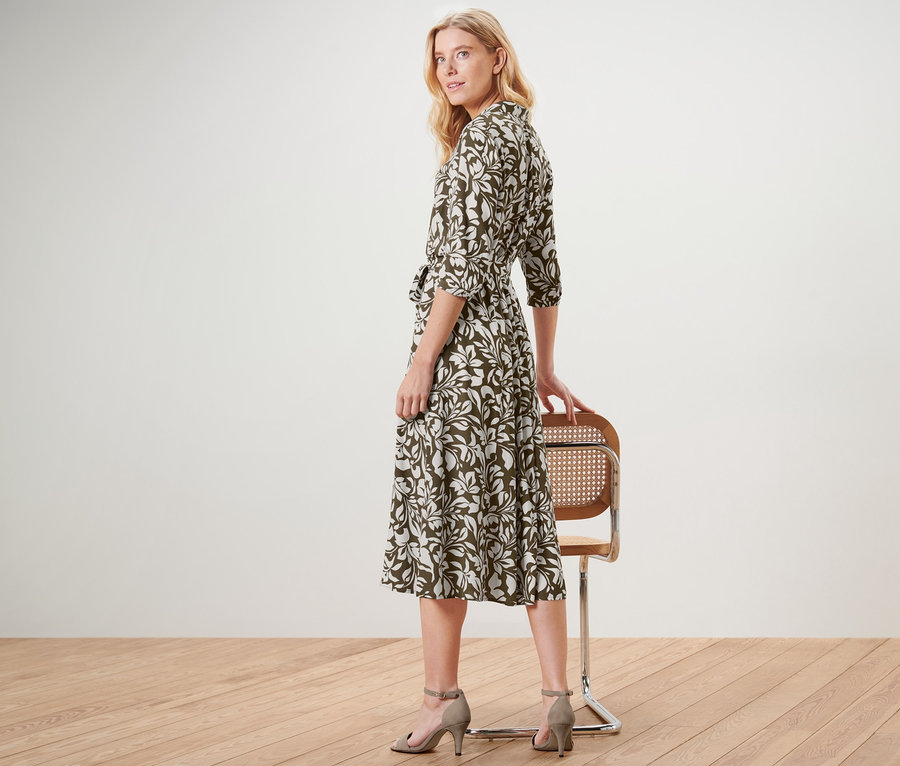Frau blickt über die Schulter und posiert in einem bedruckten Hemdblusenkleid mit Gürtel und Absatzschuhen neben einem Stuhl.