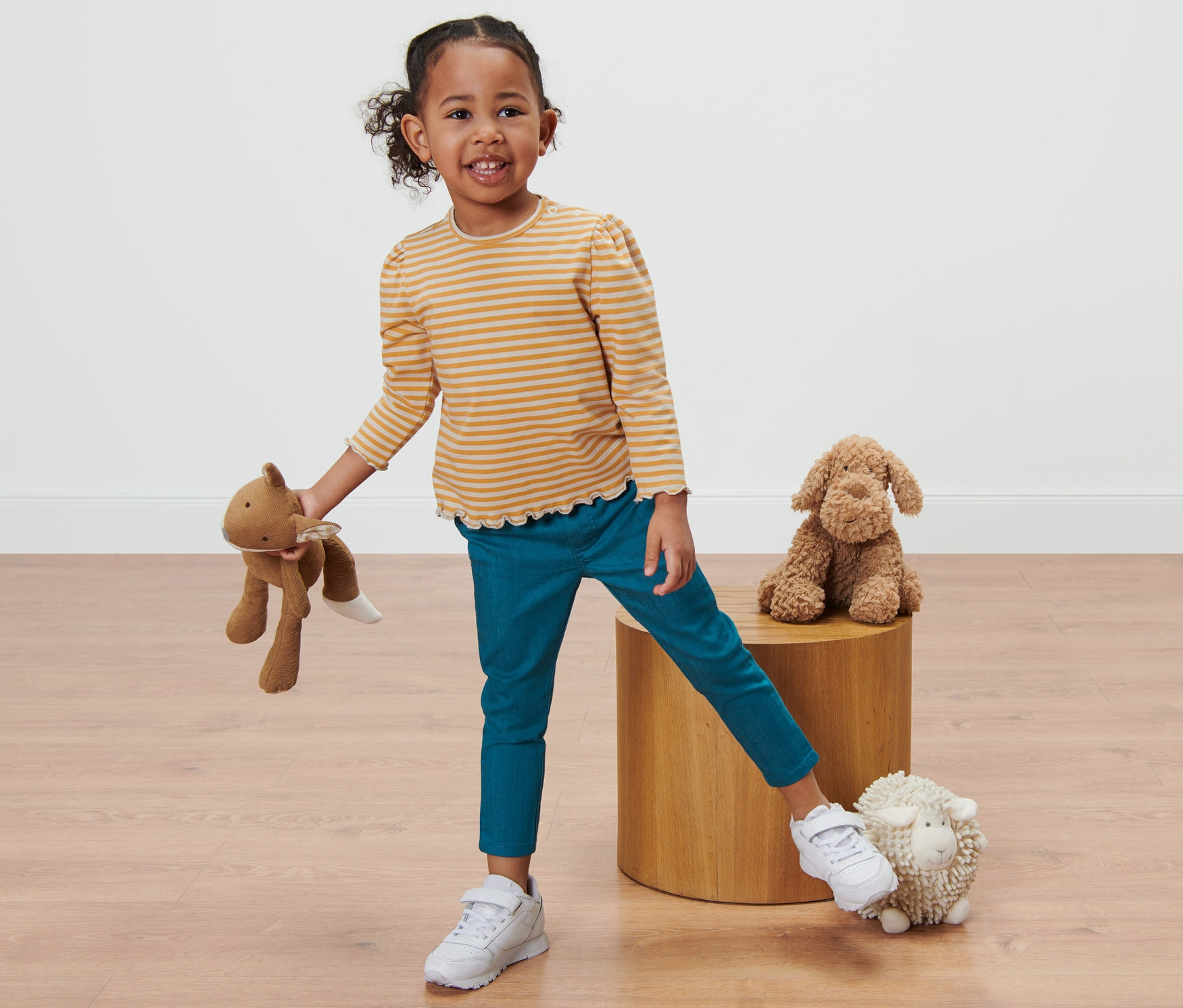 Ein lächelndes kleines Mädchen in einem gestreiften Oberteil und einer Kleinkind-Treggings posiert mit Stofftieren.