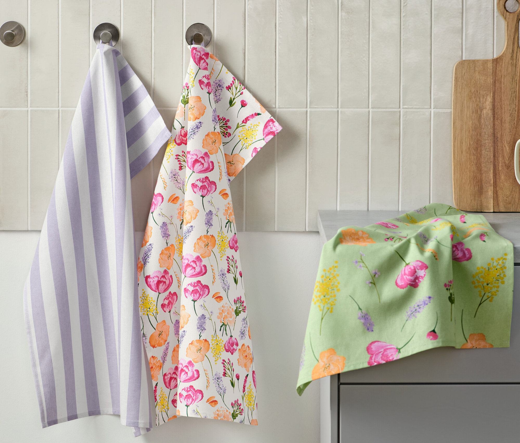 Drei Geschirrtücher hängen an der Wand: eines lila-weiß gestreift, eines mit Blumenmuster und eines liegt auf der Theke. Daneben befindet sich ein Schneidebrett und eine Tasse mit Küchenutensilien. 3 Geschirrtücher