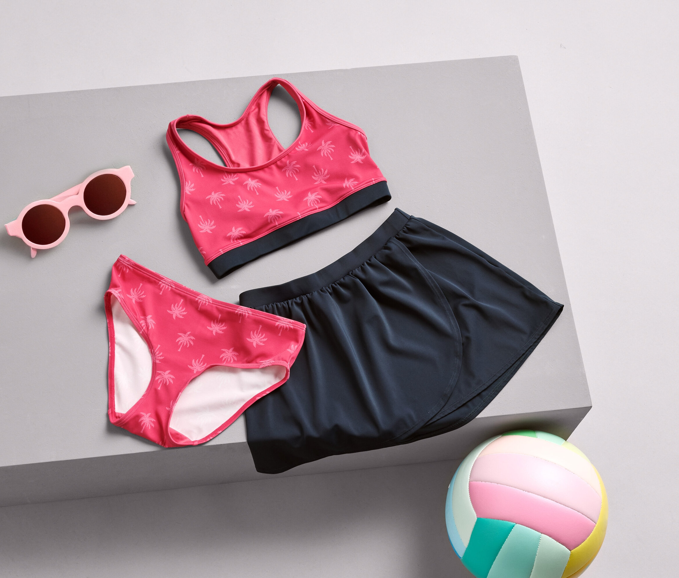 Pinke Sonnenbrille, Bikinihose mit Palmenmuster, Sport-BH und marineblauer Rock liegen auf einer grauen Oberfläche, zusammen mit einem bunten Volleyball.