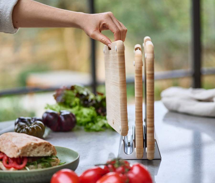 Eine Hand greift nach einem Schneidebrett in einem Halter auf einer Küchentheke, auf der sich auch ein Sandwich, Salat und Tomaten befinden.