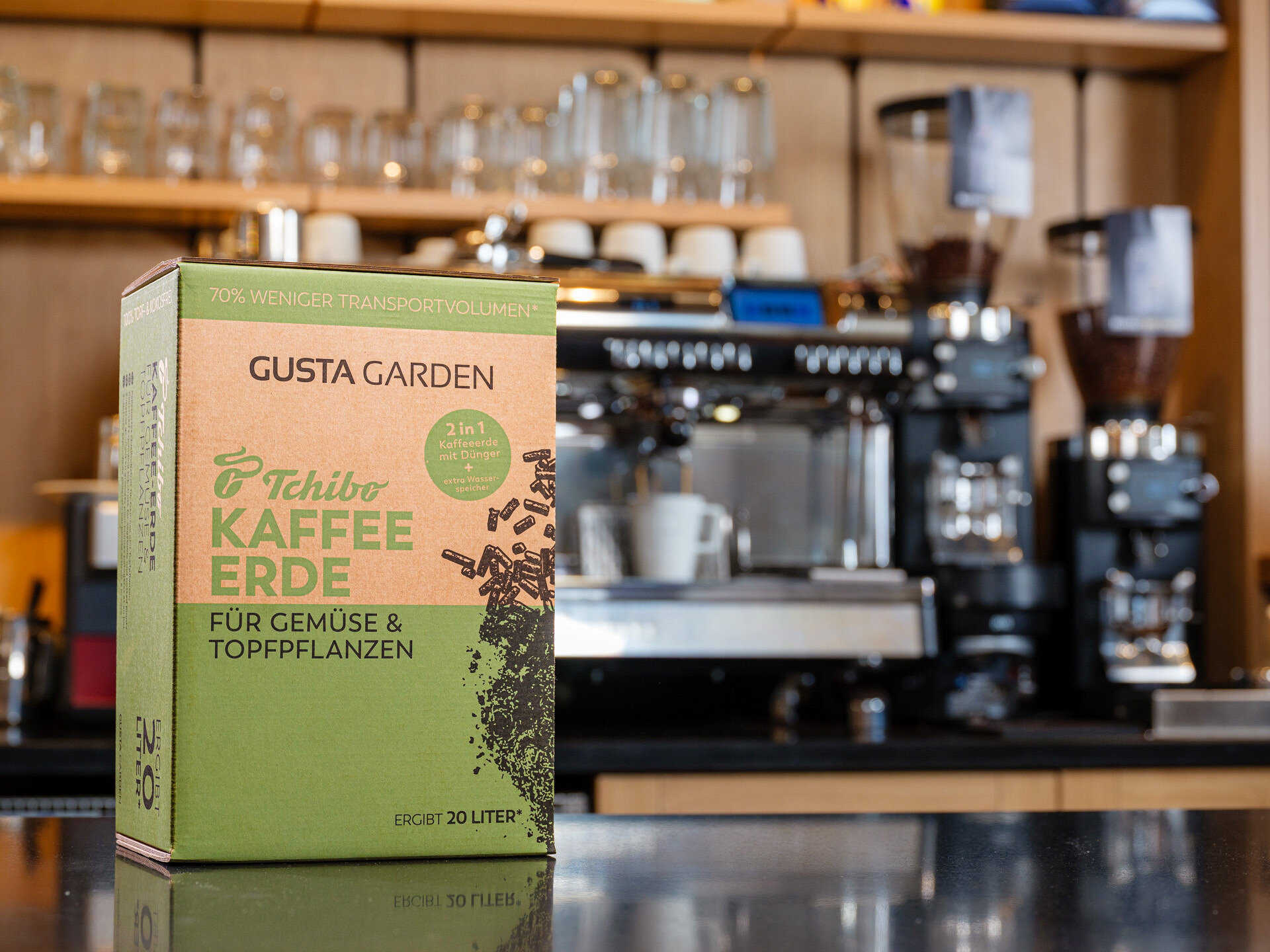 Eine Packung Tchibo Kaffee Erde steht vor einer Kaffeemaschine. Die Packung ist grün und braun und trägt die Aufschrift "Gusta Garden".