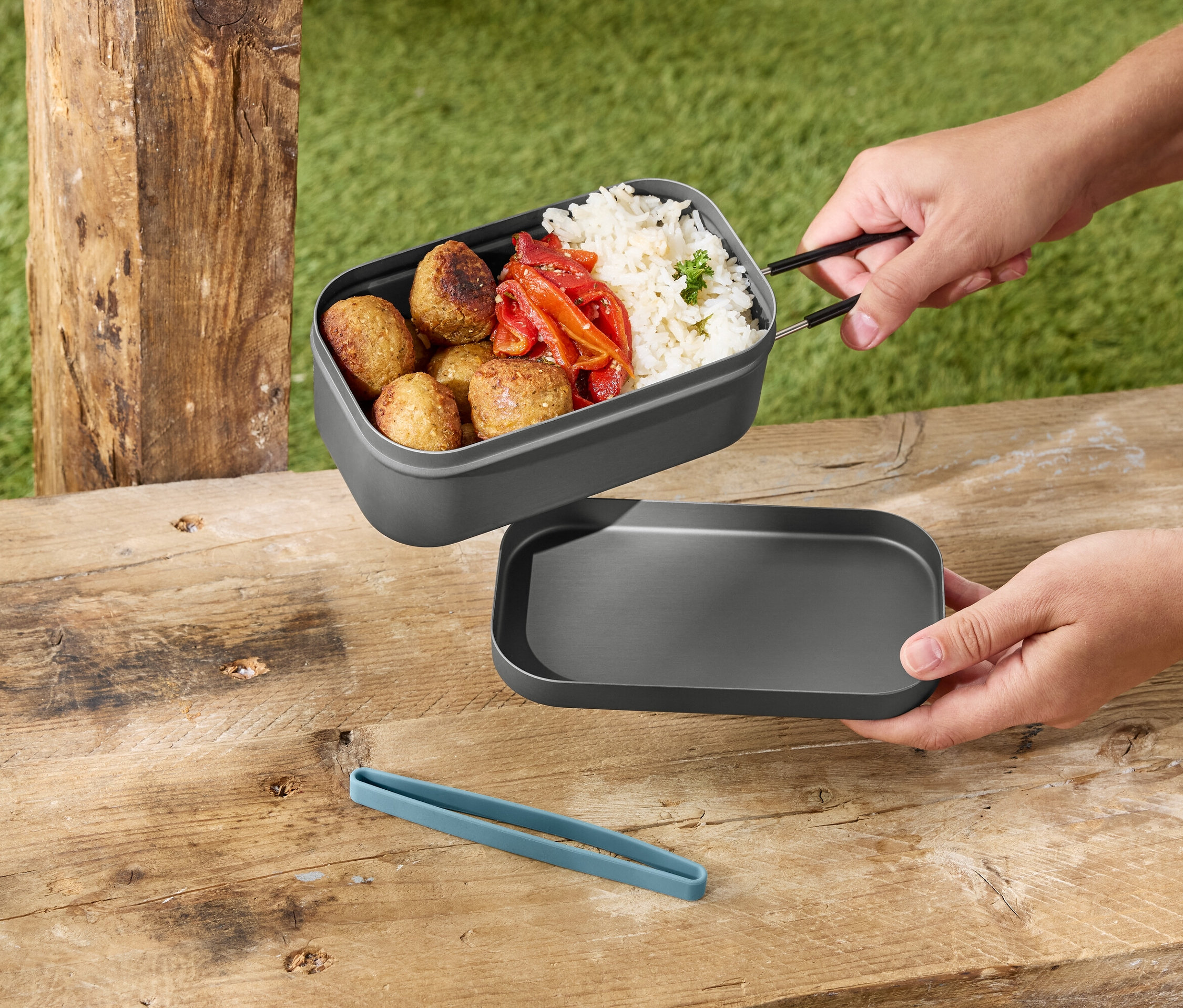 Hände halten eine graue Lunchbox mit Reis, Fleischbällchen und Paprika über einem grauen Deckel auf einem Holztisch.