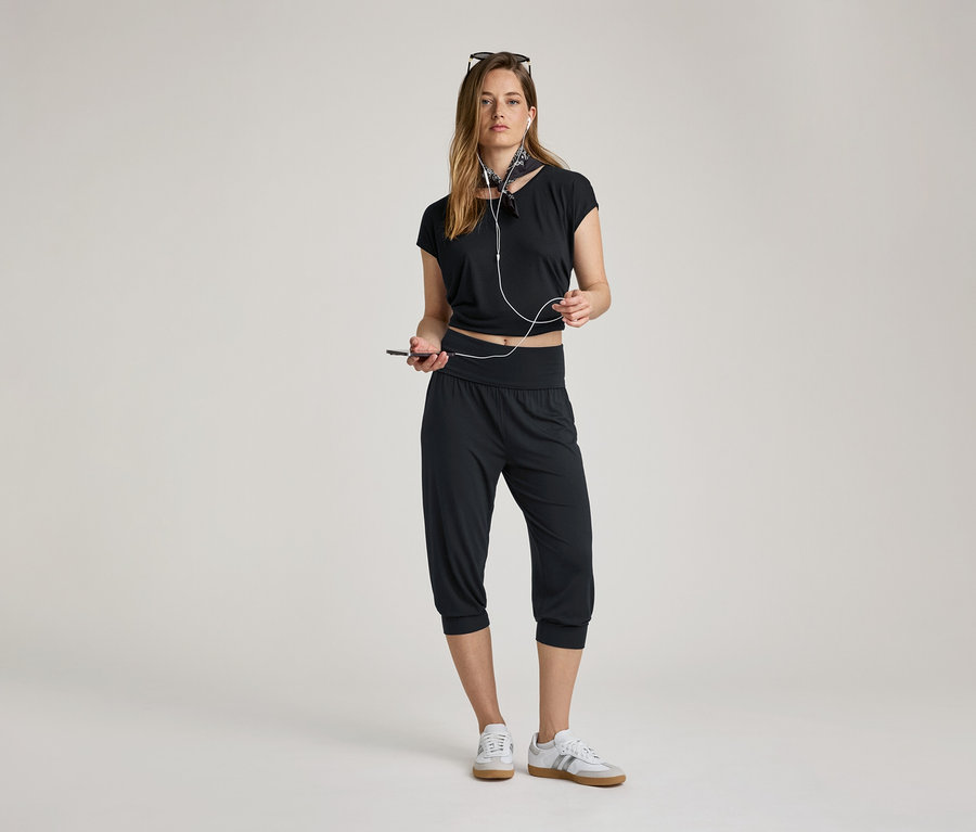 Frau mit Kopfhörern und Handy. Sie trägt eine 3/4-Yogahose, ein schwarzes Oberteil und Turnschuhe.