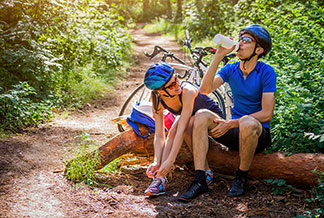Ein junges Paar, das kurze Fahrradbekleidung und Fahrradhelme trägt, sitzt auf einem Baumstamm und macht bei einer Fahrradtour eine Trinkpause im Wald.
