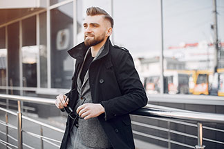 Herrenjacke fürs Büro: Ein junger Mann, der einen schwarzen Kurzmantel und darunter ein graues Sakko trägt, lehnt an einem Geländer und hält seine Sonnenbrille dabei in der Hand.
