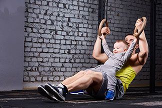 Ein junger Vater in Sportshorts hält seinen Sohn auf dem Schoß, und trainiert gleichzeitig seine Arme an Ringen im Fitnessstudio.