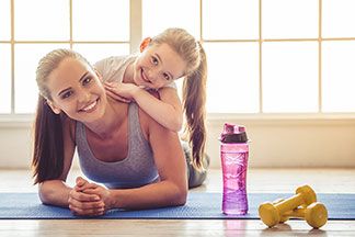 Eine junge Mutter und ihre Tochter liegen auf einer Yogamatte und lachen glücklich in die Kamera.