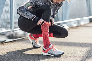 Eine junge Frau zieht sich ihre rot-schwarz gemusterten Sportsocken, die sie in den Sportschuhen trägt, bis zur Mitte des Unterschenkels hoch.