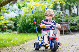 Spielzeug für draußen im richtigen Alter nutzen: Ein kleiner Junge, der kurze Hosen und ein rosa-blau-gestreiftes Shirt trägt, fährt mit seinem Dreirad über einen Plattenweg im Garten.