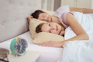 Entspannt aufwachen: Eine junge Frau liegt wach im Bett, neben ihr ein schlafender Mann und auf dem Nachttisch steht ein Wecker.