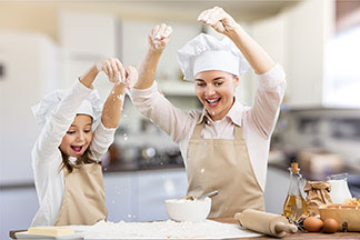 Küchentextilien zum Backen: Mutter und Tochter sind beim Backen in der Küche, tragen beide weiße Kochmützen sowie beige Schürzen und streuen mit nach oben gestreckten Händen Mehl auf ein Backblech.