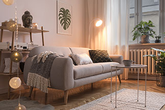 Gemütliches Ambiente durch die Wohnzimmerbeleuchtung: Eine graue Couch in einem Wohnzimmer, davor ein runder metallener Sofatisch auf einem gestreiften Teppich, dahinter eine kleine Stehlampe.
