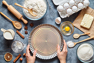 Backzubehör für Ihr Zuhause: Ein Tisch auf dem eine Kuchenform, ein Nudelholz, verschiedene Kochlöffel und Schalen, sowie die Backzutaten Eier, Mehl, Milch und Gewürze stehen.