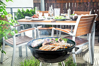 Ein Balkon, auf dem ein Grill mit Fleisch steht. Im Hintergrund ist ein rustikaler, gedeckter Gartentisch mit Holzstühlen zu sehen.