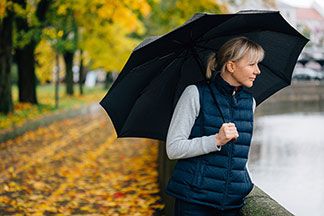Gut für den Stadtbummel: Eine blonde Frau, die eine schwarze Sportweste trägt, schlendert mit einem Regenschirm auf einem Bürgersteig an einem Fluss entlang.