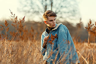 Jacke für die Freizeit: Ein junger Mann geht bei Abendstimmung durch ein herbstliches Feld, dabei trägt er seine Jeansjacke mit weißem Wollkragen über der Schulter.