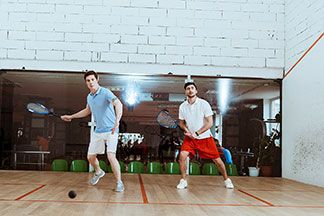 Zwei junge Männer, die beide mittellangen Sportshorts tragen, spielen in einer Halle Squash.