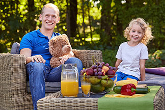 Ein Vater sitzt mit seiner kleinen Tochter auf Rattan-Gartenmöbeln – vor ihnen steht ein Rattan-Tisch, der mit einer Obstschale, Gemüse und Orangensaft gedeckter ist.