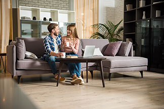 Gemütlichkeit im Wohnzimmer: Ein junges Paar sitzt auf einer fliederfarbenen Eckcouch im Wohnzimmer und unterhält sich.