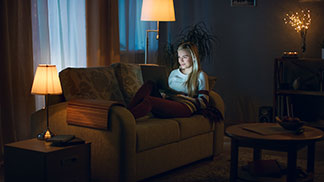 Gute Qualität für Ihre Wohnzimmerbeleuchtung: Eine junge, blonde Frau liegt auf ihrer Couch und schaut in den Laptop, während das Wohnzimmer schummrig von drei Lampen ausgeleuchtet wird.