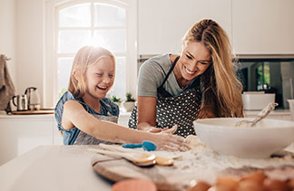 Backzubehör mit Gütesiegel: Eine junge blonde Frau und ihre Tochter stehen zusammen in der Küche und backen – beide tragen Schürzen und lachen herzhaft.