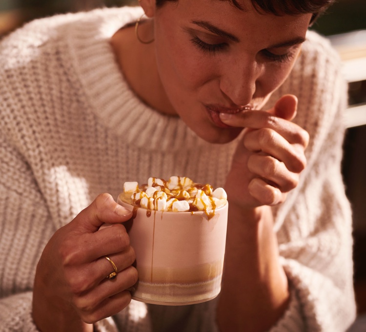Eine Frau hält eine Tasse mit einem Flavoured Coffee mit Marshmallows und Karamellsirup in der Hand