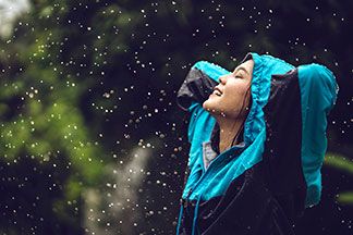 Eine junge, asiatische Frau, die eine blau-schwarze Regenjacke samt Kapuze trägt, steht im Regen, verschränkt die Arme hinter dem Kopf, und schaut mit geschlossenen Augen lachend gen Himmel.