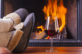 Hausschuhe für den Winter: Nahaufnahme von kuscheligen Hüttenschuhen vor einem Kamin. Daneben steht ein Glas Rotwein.