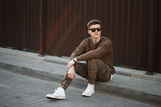 Sportschuh für den Alltag: Ein stylisch gekleideter, junger Mann, der weiße, knöchelhohe Sneaker trägt, sitzt lässig an einer Bordsteinkante.