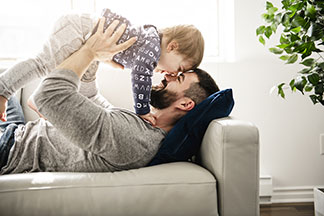 Ein Vater in grauem Langarmshirt spielt zusammen mit seinem Sohn auf der Couch.