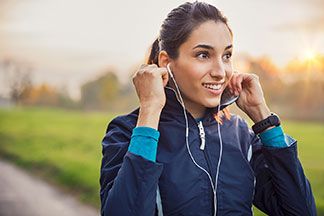 Regenjacken für den Sport: Eine junge, dunkelhaarige Frau, bekleidet mit einer blauen Softshelljacke und weißen Kopfhörern im Ohr, macht sich bereit zum Joggen in der Natur.