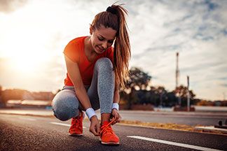 Eine junge Frau, die eine graue Laufhose und ein orangenes T-Shirt trägt, bindet sich in der Hocke ihren orangenen Sportschuh zu.