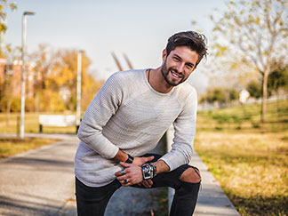 Ein junger, zufrieden lächelnder Mann mit Bart steht mit einem hellen Langarmshirt in einem Park.