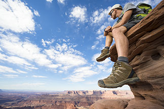 Das richtige Material für Ihren Sportschuh: Ein junger Mann in T-Shirt und kurzer Hose lässt seine Trekkingschuhe bei einer Rast auf einem Felsen nach unten baumeln.