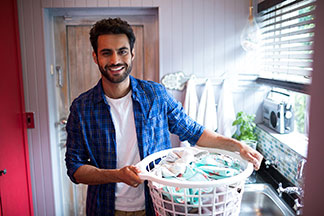 Herrenmode richtig pflegen: Ein junger Mann, der mit einem blauen, offenen Hemd und einem weißen T-Shirt bekleidet ist, trägt einen weißen, runden Plastikkorb voller Wäsche.