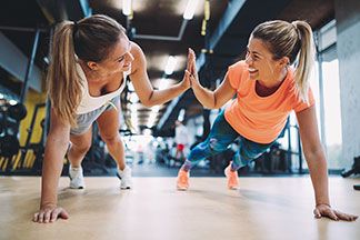 Zwei junge Frauen trainieren im Fitnessstudio, beide stehen in Liegestütz mit einer Hand auf dem Boden und mit der anderen Hand klatschen sie sich ab.