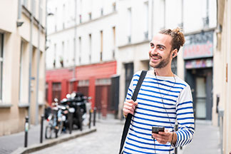 Ein fröhlicher, junger Mann in einem blau-weiß geringelten Langarmshirt spaziert mit einem Smartphone in der Hand durch eine Stadt.