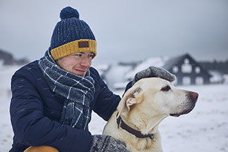 Das ideale Accessoire für den Winter: Ein junger Mann, der Schal, Mütze und Handschuhen trägt, streichelt in der Hocke seinen weißen Labrador in einer verschneiten Winterlandschaft.