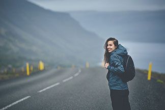 Gut ausgerüstet: Eine junge Frau, die eine Regenjacke und einen Rucksack trägt, läuft an einem regnerischen Tag auf einer verlassenen Straße durch die Natur.