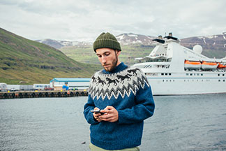 Ein junger, bärtiger Mann steht in einem Norwegerpullover vor einem Hafen, wo gerade ein Passagierschiff einläuft, und schaut auf sein Smartphone.