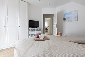 Zeitloses Design: In einem hell eingerichteten Schlafzimmer steht ein weißer Schlafzimmerschrank mit drei Türen sowie eine Kommode mit grau-weißen Schubladen, auf der ein Fernseher steht.